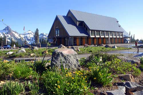 A modern wooden building with a steep roof stands surrounded by wildflowers and rocky terrain. Snow-capped mountains and a clear blue sky are in the background. Nearby, a person walks along a pathway.