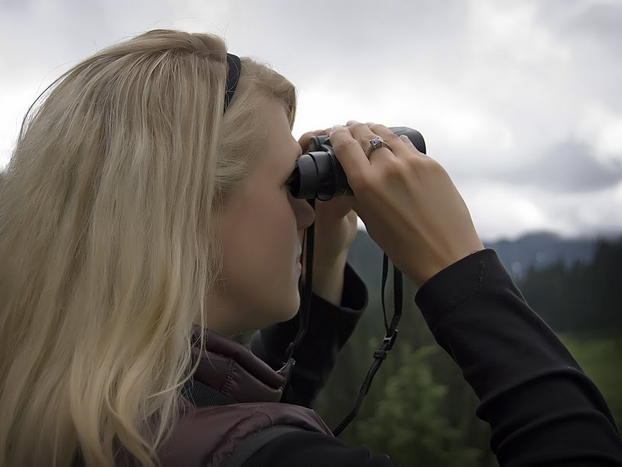 A woman with long blonde hair looks through binoculars, standing outdoors with a cloudy sky and forested hills in the background. She wears a dark jacket and a headband.