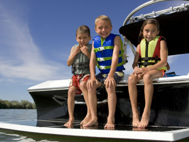 Three children in life jackets sit on the edge of a boat, smiling at the camera. The sunny day and calm water create a cheerful, relaxing atmosphere.