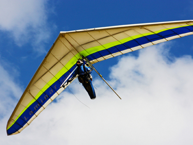 A person hang gliding through a clear blue sky with white clouds, supported by a wing with blue and green stripes.