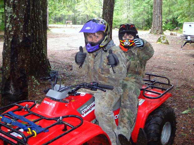 Two people wearing helmets and camouflage clothing sit on a red ATV in a forested area. Both give thumbs up, with tall trees surrounding them and a glimpse of a trailer or building in the background.