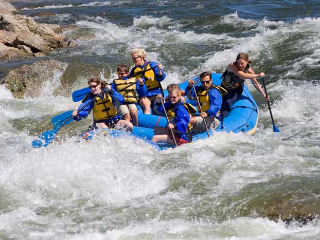 A group of people in life jackets are white-water rafting in a blue inflatable raft, navigating through turbulent waters and waves. They are holding paddles and appear to be focused on steering the raft through the rapids.