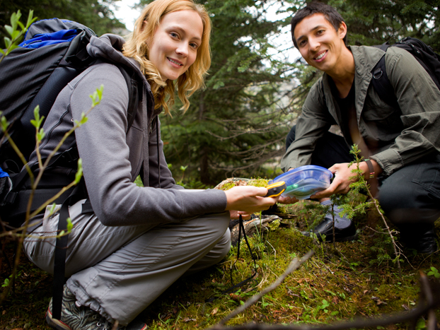 Two people outdoors in a forest, crouching on the ground. They are holding a small container and appear to be engaged in a recreational activity, possibly geocaching or studying nature. Both are smiling, with backpacks and wearing casual outdoor clothing.