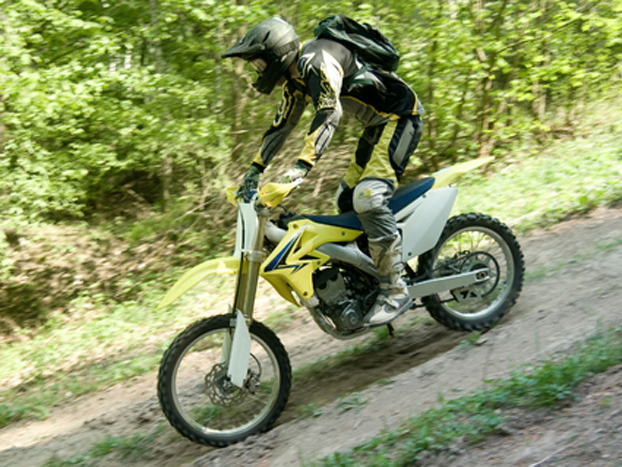 A person wearing protective gear rides a yellow dirt bike up a forested trail, surrounded by green trees on a sunny day.